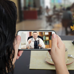 Woman sits at a cafe table and communicates with telemedicine doctor by cellphone. In touchscreen, male physician reviewing brain x-ray image. Patient has ability to reach telemedicine specialist remotely by mobile application. Horizontal indoors back side shot on cafe blurred background